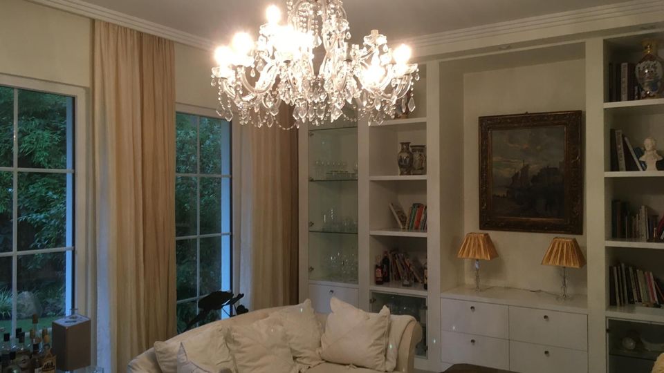 Elegantes Wohnzimmer mit großem Kristalllüster, hellem Sofa und maßgefertigten weißen Einbauregalen mit Vitrinen und Schubladen. Große Fenster mit bodenlangen Vorhängen geben den Blick ins Grüne frei; an der Regalwand hängen ein gerahmtes Bild und stehen zwei Tischlampen.