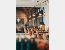 Ein modernes Restaurant mit eleganten, gepolsterten Stühlen in warmen Farbtönen und gedeckten Tischen. Im Hintergrund ist eine Wand mit einem großformatigen Foto von einem historischen, bogenförmigen Kircheninterieur zu sehen, beleuchtet durch hängende Glühbirnenlampen.