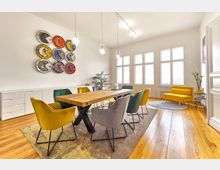 Heller Besprechungsraum mit großem Holztisch, umgeben von Stühlen in unterschiedlichen Farben. An der Wand hängt eine farbige Wandkunst aus kreisförmigen Elementen, die an Pac-Man-Motive erinnern; der Raum hat Holzboden, gelbe Sitzmöbel und große Fenster, die viel Tageslicht hereinlassen.