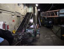 Industriehalle mit Arbeitsgeräten und einer Metalltreppe, die zu einem oberen Bereich führt. Im Hintergrund stehen eine grüne Lokomotive und verschiedene Werkzeuge sowie Bauteile, während Kabel und Maschinen im Vordergrund verteilt sind.