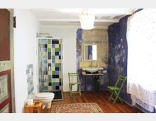 Rustikal eingerichtetes Badezimmer mit Holzboden und einer gläsernen Dusche mit farbigen Fliesen in Blau-, Grün- und Gelbtönen. Links befindet sich eine Toilette, daneben ein Waschbecken auf einem antiken Tischgestell vor einer blau und gelb gestalteten Wand, ergänzt durch einfache grüne Holzstühle und einen gemusterten Teppich.