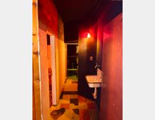 Ein schmaler Innenraum mit Holz- und dunklen Wandverkleidungen, beleuchtet von rotem Licht. Im Vordergrund hängt ein kleines Waschbecken an der Wand, während der Boden aus geflickten, bunten Fliesen besteht.