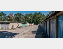 Reihe von Garagen mit Holz- und Metalltoren entlang eines betonierten Platzes, umgeben von Bäumen in einem Waldgebiet. Im Hintergrund sind vereinzelte Straßenlaternen sichtbar.