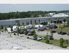 Industriekomplex mit flachen, grauen Gebäuden und einem großen Parkplatz, auf dem viele Autos sowie einige Wohnmobile und Transporter geparkt sind. Im Hintergrund sind Bäume und weitere Gebäude zu sehen.