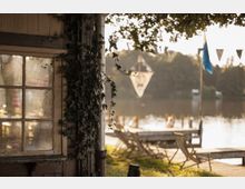 Ein Uferbereich mit einem Holzgebäude im Vordergrund, dessen Fenster von Efeu bewachsen ist. Im Hintergrund befinden sich ein Steg und Liegestühle am Wasser, eingerahmt von Bäumen und einer ruhigen Seeoberfläche.