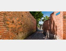 Schmale Kopfsteinpflastergasse zwischen hohen roten Backsteinmauern, die in Richtung kleiner Gebäude und eines Eingangstores führt. Oben rechts ist ein Straßenschild „Glintzmauer“ zu sehen, darüber ragen Bäume in den Himmel.