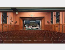 Theaterraum mit einer großen Bühne im Hintergrund, eingerahmt von Holzvertäfelungen und einer roten Wand mit blauen Akzentstreifen. Im Vordergrund sind Reihen von leeren Holzstühlen zu sehen, während auf der Bühne Kulissen und Theaterrequisiten aufgebaut sind.
