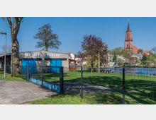 Moderner Vereinsbereich am Wasser mit einem Gebäude der "Sektion Rudern" und einer umzäunten Grünfläche. Im Hintergrund ist eine Kirche mit rotem Backsteinturm sowie weitere Häuser und Bäume zu sehen.