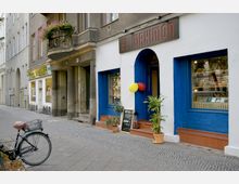 Straßenansicht mit einem kleinen Ladenlokal namens "El Maximón", das einen weiß-blauen Anstrich hat. Vor der Eingangstür sind Pflanzen in Töpfen, bunte Luftballons und ein Schild aufgestellt, während ein Fahrrad am Gehweg lehnt.