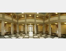 Ein Innenraum mit einer zweistöckigen Halle, gekennzeichnet durch Säulen aus hellem Stein, ein Schachbrettmuster auf dem Boden und mehrere Türen im Erdgeschoss. Die Decke ist gewölbt und mit runden Deckenleuchten beleuchtet.