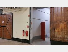Ein Industriegebäude-Innenraum mit Holztoren, einer weiß gestrichenen Backsteinwand, einem roten Türrahmen mit offenstehender Tür und zwei an der Wand befestigten Feuerlöschern. Sicherheitskennzeichnungen und sichtbare Rohrleitungen ergänzen die Szene.