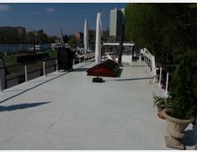 Oberdeck eines Bootes mit weißem Bodenbelag, umlaufendem Geländer und vereinzelten Pflanzen in Töpfen. Im Hintergrund sind ein Fluss, weitere Boote, Bäume sowie Wohn- und Hochhäuser sichtbar.