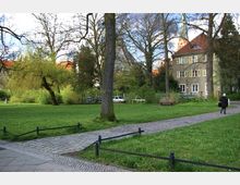 Ein kleiner Park mit Grünflächen, Bäumen und einem gepflasterten Weg, der an einer Bank vorbeiführt. Im Hintergrund stehen Wohnhäuser mit roten Dächern, teils verdeckt durch Bäume.