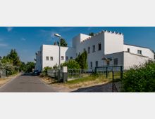 Weiße Gebäude mit flachen Dächern und zinnenartigen Verzierungen entlang der Dachkante, daneben ein Garten mit Zaun und Gehweg. Umgebung mit wenigen Bäumen und einer schmalen Straße unter einem blauen Himmel.