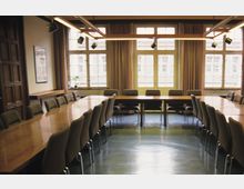 Konferenzraum mit großen, rechteckigen Holztischen, die in U-Form angeordnet sind, und gepolsterten Stühlen. Der Raum hat große Fenster mit Vorhängen, die viel Tageslicht hineinlassen, sowie Deckenlampen und beige Wände.