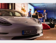 Ein weißes Tesla-Auto steht im Vordergrund eines Innenraums mit einer Bühne im Hintergrund, auf der zwei Personen in einem Vortrag oder einer Präsentation stehen. Im Raum befinden sich Beleuchtungen, ein Bildschirm, Stühle und weitere Veranstaltungsrequisiten.