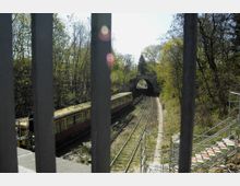 Blick durch Metallgitter auf eine zweigleisige Bahnstrecke in einem bewaldeten Gebiet, mit einem gelb-roten Zug auf dem linken Gleis und einer kleinen Brücke im Hintergrund. Rechts führt eine Treppe aus Metallgerüsten zum Bahndamm.