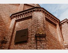 Rote Backsteinfassade eines Gebäudes mit dekorativen Details, darunter ein goldfarbenes Ornament und ein Schild mit der Aufschrift „21 gramm“. Im Hintergrund ein blauer Himmel.