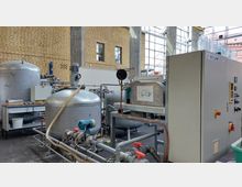 Innenraum einer industriellen Anlage mit verschiedenen Edelstahlbehältern, Rohrleitungen und einer Steuerungseinheit. Im Hintergrund sind große Fenster und eine Wand aus gelben Klinkersteinen sichtbar, die viel Tageslicht in den Raum lassen.