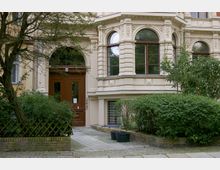 Historische Fassade eines Wohngebäudes mit verzierten Fenstern und einem braunen Holzeingang. Vor dem Gebäude befinden sich ein gepflasterter Weg, Büsche und ein Baum.