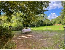 Grüne Wiese mit vereinzeltem Baumbestand und einem Tisch mit weißer Tischdecke und Bänken im Vordergrund. Im Hintergrund ist eine offene Rasenfläche, umgeben von Bäumen, zu sehen.