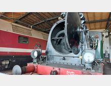 Eine geöffnete Dampfmaschine in einer Lokhalle, die den Blick auf das Innenleben des Kessels erlaubt. Im Hintergrund steht eine rot-weiße Diesellokomotive, und das Gebäude zeigt ein Holzdach mit Metallträgern.