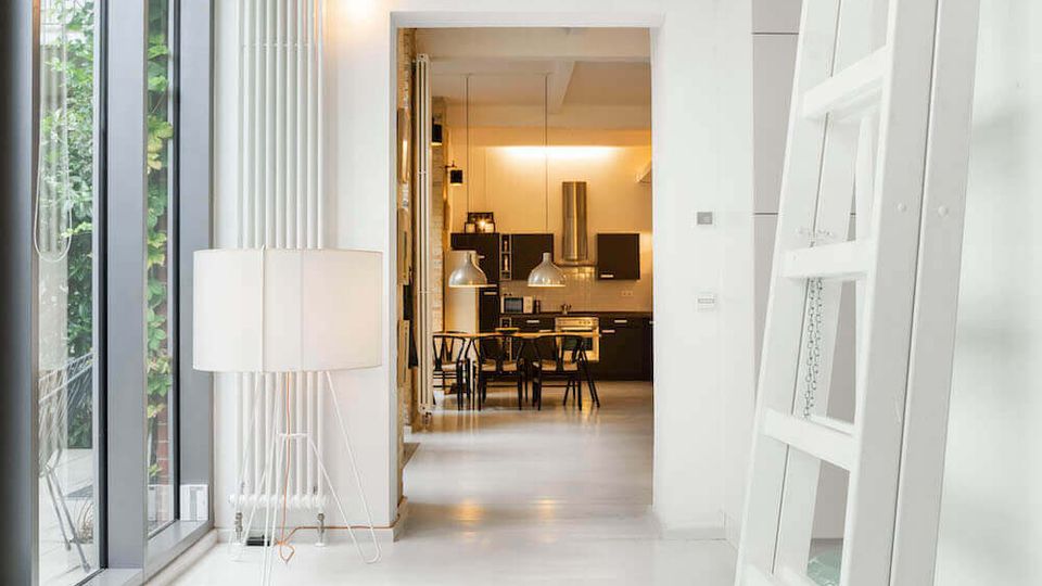 Bright modern hallway with white floors and walls, a tall window wall on the left, and a leaning white ladder on the right. The corridor leads through an open doorway to a warmly lit kitchen and dining area with pendant lights and dark cabinetry.