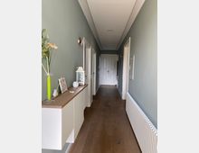 Ein schmaler, langer Flur mit hellgrünen Wänden, weißen Zimmertüren und dunklem Holzfußboden. Links steht ein weißes Sideboard mit Dekorationen, rechts verläuft ein langer Heizkörper, und am Ende des Flurs befindet sich eine weiße Tür mit goldfarbener Klinke.