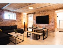 Gemütliches Wohnzimmer mit freiliegenden Backsteinwänden, dunklem Sofa, modernem Esstisch mit Holzbänken und gedecktem Geschirr. An der Wand hängt ein großer Flachbildfernseher, und das Licht fällt durch ein kleines Fenster mit einer Pflanze.