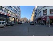 Straßenansicht einer städtischen Umgebung mit mehrstöckigen Gebäuden auf beiden Seiten, darunter ein modernes Geschäftshaus links mit Apothekenbeschilderung und ein älteres Gebäude rechts mit kleinen Läden und Restaurants. Geparkte Autos säumen die Straße, und eine Person geht mittig in Richtung eines Hintergrunds mit Bäumen und weiteren Gebäuden.