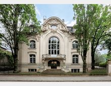 Historische Stadtvilla im Jugendstil mit aufwendig verzierten Fassadendetails und großem zentralen Bogenfenster, flankiert von Bäumen auf beiden Seiten. Ein schmiedeeisernes Zauntor umgibt das Gebäude, das eine symmetrische Architektur aufweist.