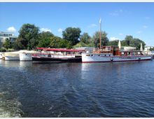 Boote liegen an einem Kai auf einem Fluss, umgeben von grünen Bäumen und einigen Gebäuden im Hintergrund. Der Himmel ist klar und blau, und das Wasser spiegelt die Boote und die Umgebung wider.