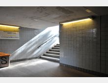 Unterführung mit grauen, gefliesten Wänden und einer Treppe, die ins Tageslicht führt. An der Wand ist ein gelbes Wegweiser-Schild mit Richtungsangaben zu nahegelegenen Straßen und Stationen befestigt.