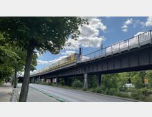 Eine gelbe U-Bahn fährt auf einer erhöhten Stahlbrücke über eine Straße, die von Bäumen gesäumt ist. Im Hintergrund sind grüne Vegetation und ein teilweise bewölkter Himmel zu sehen.