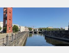 Ein urbaner Kanal mit steinernen Ufermauern, flankiert von einem roten Backsteinturm und modernen Gebäuden auf der linken und rechten Seite. Im Hintergrund sind eine Brücke mit Statuen, umgebende Grünflächen und Baustellenkräne sichtbar unter klarem Himmel.
