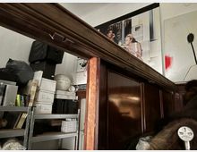 Innenraum eines Lager- oder Atelierraums mit hohen, dunkel vertäfelten Holzelementen und einem Metallregal voller Kisten, Taschen und Materialien. An der weißen Wand hängen große Fotodrucke und ein abstraktes Bild mit rotem Farbfleck und dunkler Blütenform.