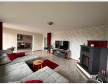 Modernes Wohnzimmer mit grauem Ecksofa, dekoriert mit roten Kissen, einem Holztisch und einem roten Teppich. Rechts im Raum steht ein dunkelgrauer Kamin, links sind Pflanzen sichtbar, und im Hintergrund öffnet sich ein Essbereich mit gemauerter Ziegelwand.