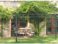 Kleiner Garten mit Rasenfläche und einer Steinmauer im Hintergrund, an der eine metallene Pergola mit Kletterpflanzen und Blättern bewachsen ist. Darunter stehen ein runder Tisch und vier Stühle aus Metall und Holz.