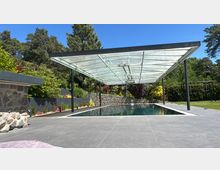 Ein moderner Swimmingpool mit einer offenen Überdachung aus Glas und Metall, umgeben von einer gepflasterten Terrasse, Steinmauern und dichter Gartenvegetation. Im Hintergrund sind Bäume und ein blauer Himmel zu sehen.