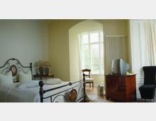 Helles Schlafzimmer mit einem schmiedeeisernen Bett, großen Fenstern mit Blick auf eine grüne Landschaft, einem antiken Holztisch mit einem Röhrenfernseher und einem gepolsterten Stuhl. Die Wände sind in einem warmen Beige gehalten, und der Boden hat einen Holzbodenbelag.