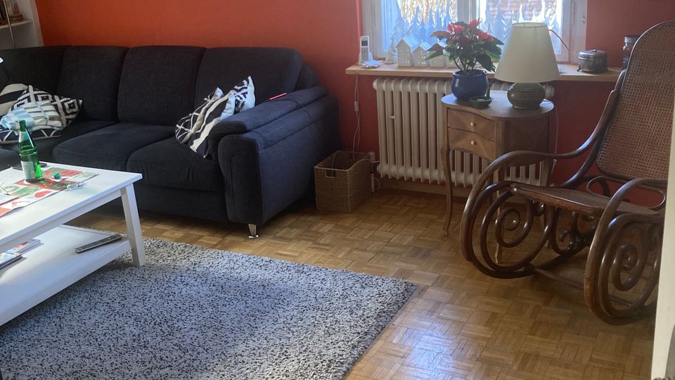 Wohnzimmer mit orangener Wand, einem dunkelblauen Sofa, einem weißen Couchtisch und einem grauen Teppich auf einem Parkettboden. Ein Fenster mit Vorhang lässt Tageslicht herein, daneben stehen ein kleiner Holztisch mit Dekorationen und ein Holz-Schaukelstuhl.