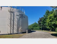 Großer, weißer Heizöltank mit der Aufschrift „Heizöl Gefahrklasse A III, 800 m³“ und einer außen angebrachten Leiter. Im Hintergrund sind ein gepflasterter Weg, grüne Bäume und blauer Himmel zu sehen.