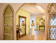 Eleganter Innenraum mit hohen Rundbögen, hellgrünen Wänden und Holzboden, flankiert von großen gotischen Türen mit Rautenglas. Im Raum stehen verzierte Goldspiegel, ein Konsolentisch, ein Kerzenleuchter und ein Kristalllüster; im Hintergrund sind große Fenster mit Sprossen zu sehen.