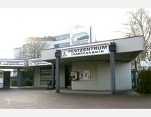 Eingangsbereich des Festzentrums Trabrennbahn mit einem Flachdach und mehreren Schildern, darunter große Schriftzüge mit der Bezeichnung des Zentrums. Im Hintergrund ist ein modernes Gebäude mit Glasfassade und weiteren Schildern zu sehen, umgeben von kahlen Bäumen.