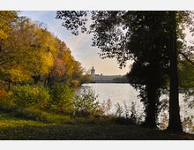 Herbstliche Parklandschaft mit Blick auf das Schloss Charlottenburg in Berlin, umgeben von Bäumen mit gelbem und braunem Laub, einem See im Vordergrund und dem Schloss im Hintergrund unter klarem Himmel.