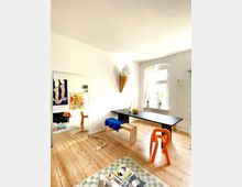 Helles Wohnzimmer mit Holzdielenboden, einem schwarzen Tisch mit Holzbank und orangefarbenem Stuhl, dekoriert mit moderner Kunst an den Wänden. Ein großes Fenster mit halbtransparenten Vorhängen lässt Tageslicht herein, während ein Spiegel und ungewöhnliche Designelemente, wie eine Wandleuchte in Form einer Eiscreme, den Raum akzentuieren.