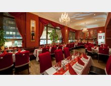 Elegantes Restaurant mit festlicher Dekoration, roten Wänden und Vorhängen sowie Kronleuchtern an der Decke. Die Tische sind mit weißen Tischdecken, roten Servietten und Weihnachtsdekoration gedeckt; die Stühle sind mit roten Hussen überzogen.