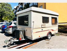 Ein älterer Wohnwagen mit beiger Außenverkleidung und schwarzen Fenstern steht auf einem Parkplatz neben geparkten Autos. Im Hintergrund sind mehrstöckige Wohnhäuser mit Balkonen und ein Baum erkennbar.