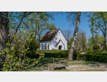 Eine kleine weiße Kapelle mit Satteldach und Holzeingang befindet sich inmitten eines grünen, baumbestandenen Friedhofs. Im Vordergrund stehen Bäume, Büsche und eine Wasserentnahmestelle.