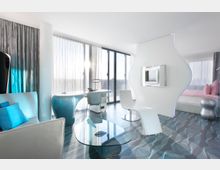 Helles, modernes Hotelzimmer mit stilvollem Design, geprägt von geschwungenen Möbeln, einem Raumteiler mit eingebautem Fernseher und dekorativen Glasoberflächen. Große Fenster mit halbtransparenten Vorhängen lassen viel Tageslicht in den Raum, während ein gemustertes Bodenmuster den futuristischen Stil betont.